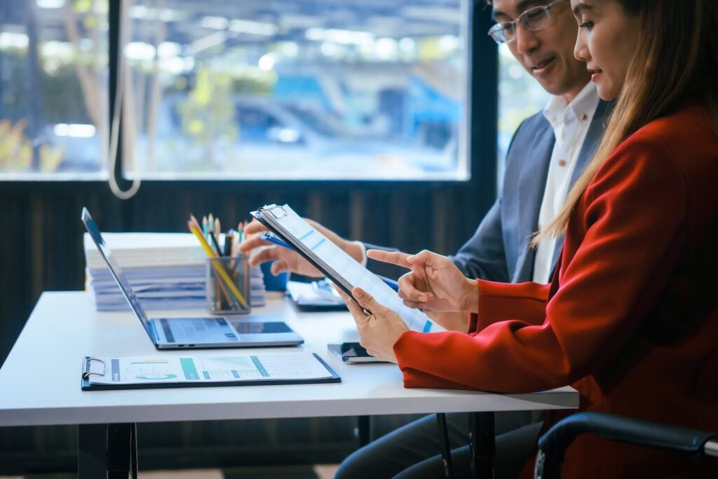Two people in business attire sit at a desk with laptops, documents, and stationery, discussing a report on indirect costs in manufacturing cost accounting in a bright office with large windows in the background.