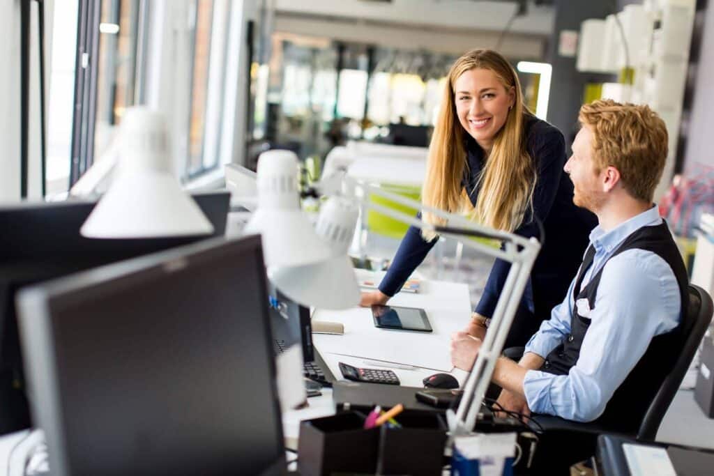 A woman stands and smiles while talking to a seated man at a modern office desk with computers, lamps, and office supplies around them. They appear to be having a collaborative discussion.