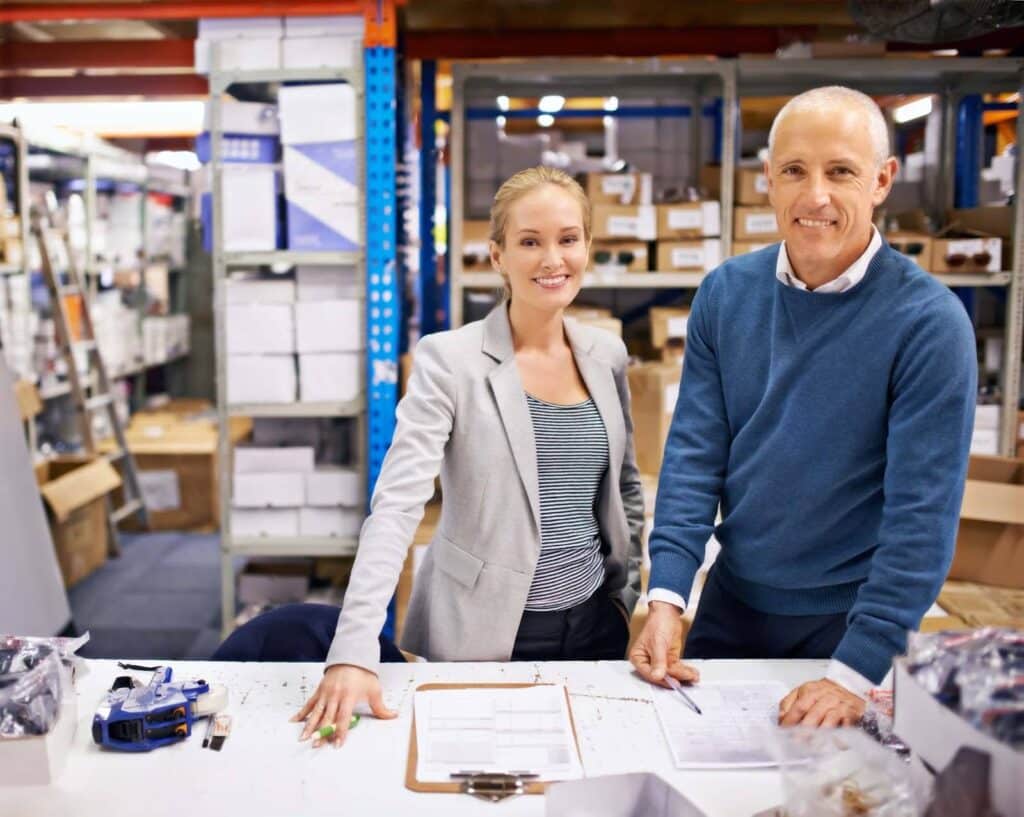 Two people, a woman in a blazer and a man in a blue sweater, stand smiling at a table in a warehouse office, surrounded by paperwork, boxes, and equipment as they discuss inventory valuation.