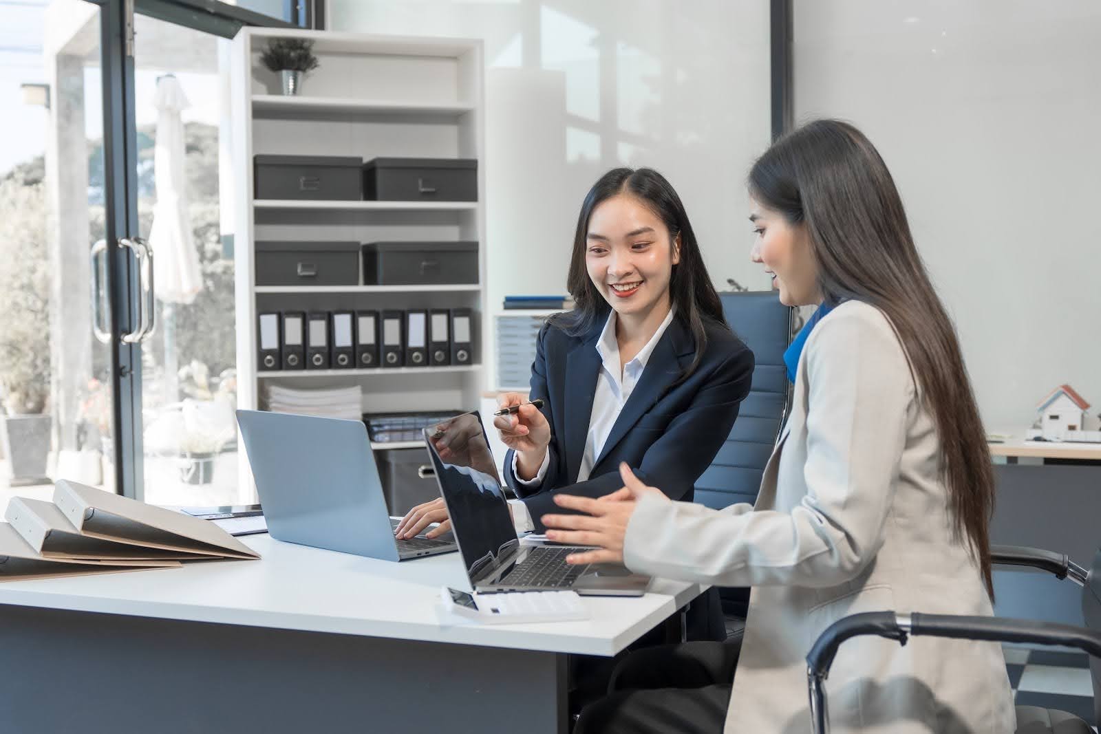 Two women in business attire sit at a desk with laptops in a modern office, smiling and discussing manufacturing cost accounting. One woman points at the other's screen as they collaborate on understanding indirect costs for their project.