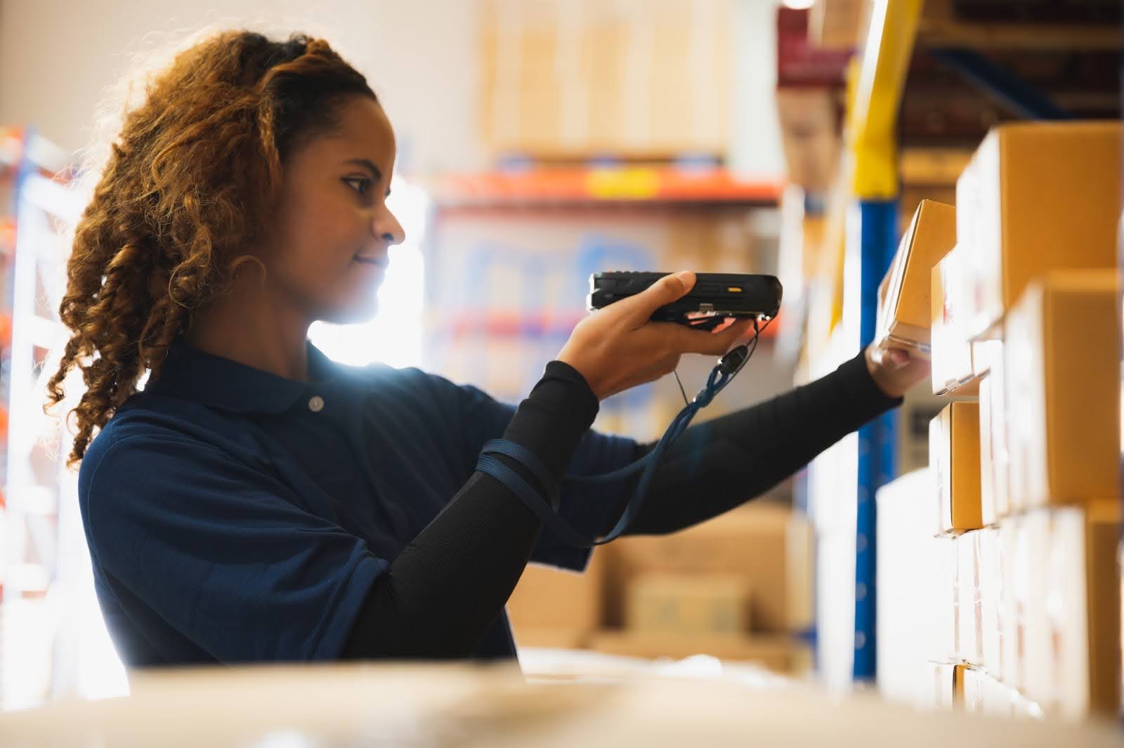 A woman wearing a navy shirt scans a package with a handheld barcode scanner while standing by shelves of boxes in a warehouse as an example of inventory management in manufacturing.