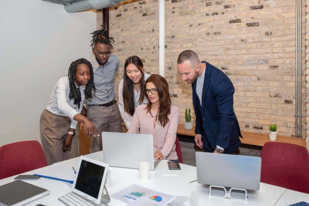 Five professionals gather around a laptop in an office, collaborating on a project. Papers, tablets, and other laptops are on the table; a brick wall and whiteboard are in the background.