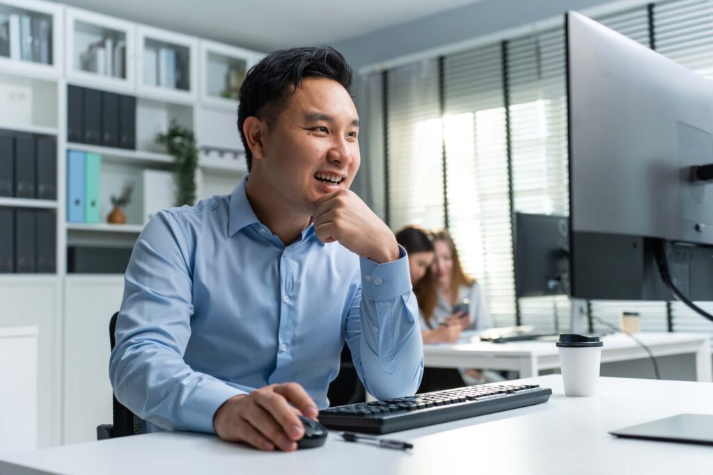 A man in a light blue shirt sits at a desk, smiling while using the Sage 100 work order module on his computer. His hand rests on a mouse beside a keyboard and coffee cup, working on Sage 100 work order module.