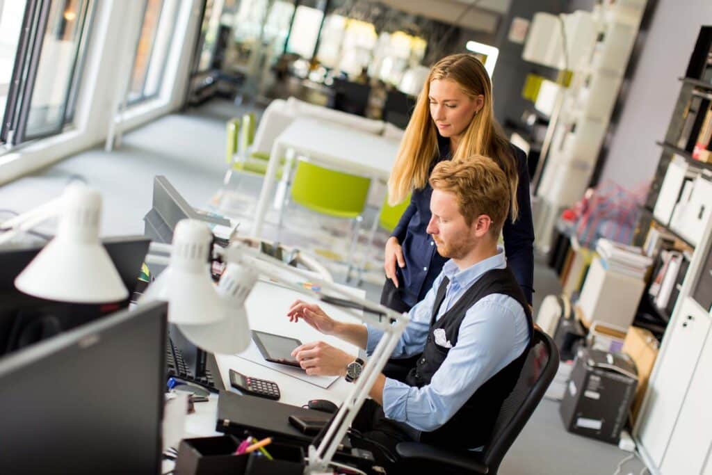 Two colleagues in a modern office, one sitting at a desk using a computer and tablet, while the other stands nearby, both focused on the screen with desks, lamps, and office supplies around them.