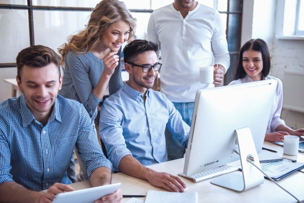 Four people smile and look at a computer screen together in a bright office, collaborating on a Sage 100 work order module. One person holds a tablet, another a cup, with papers and coffee cups scattered on the desk as they work together.