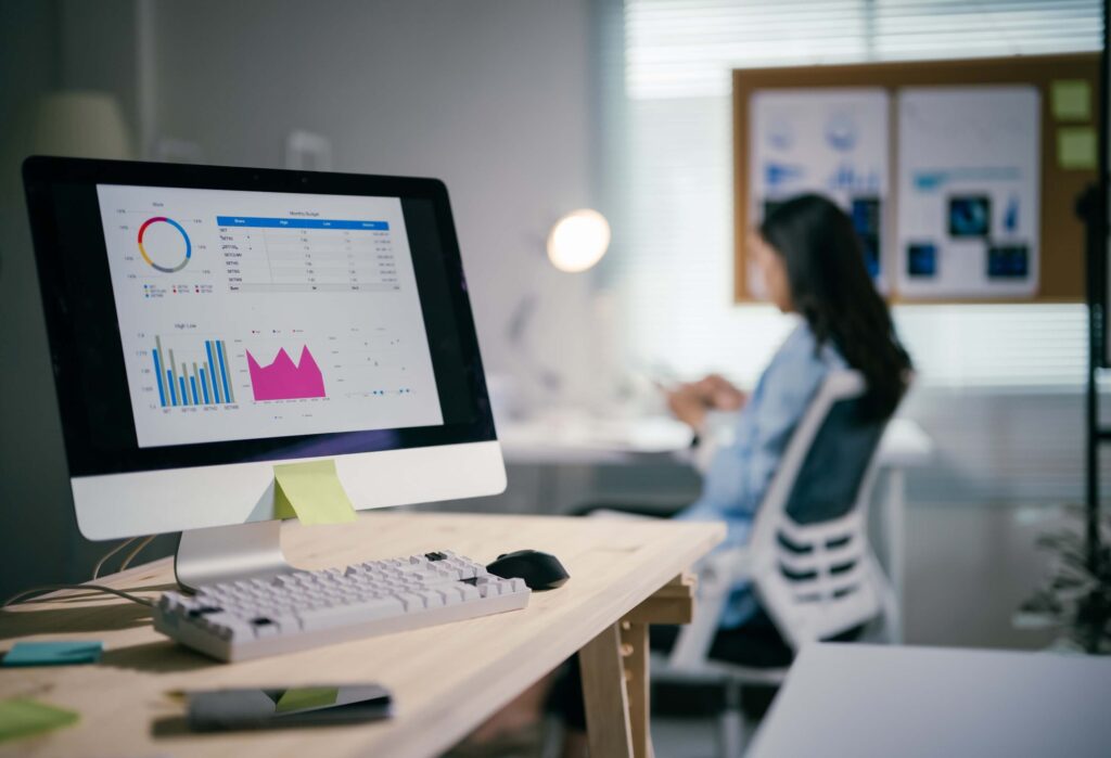 A computer monitor on a desk displays graphs and charts, with a keyboard and phone nearby. In the background, a woman sits at a desk, slightly out of focus, in a bright office setting.