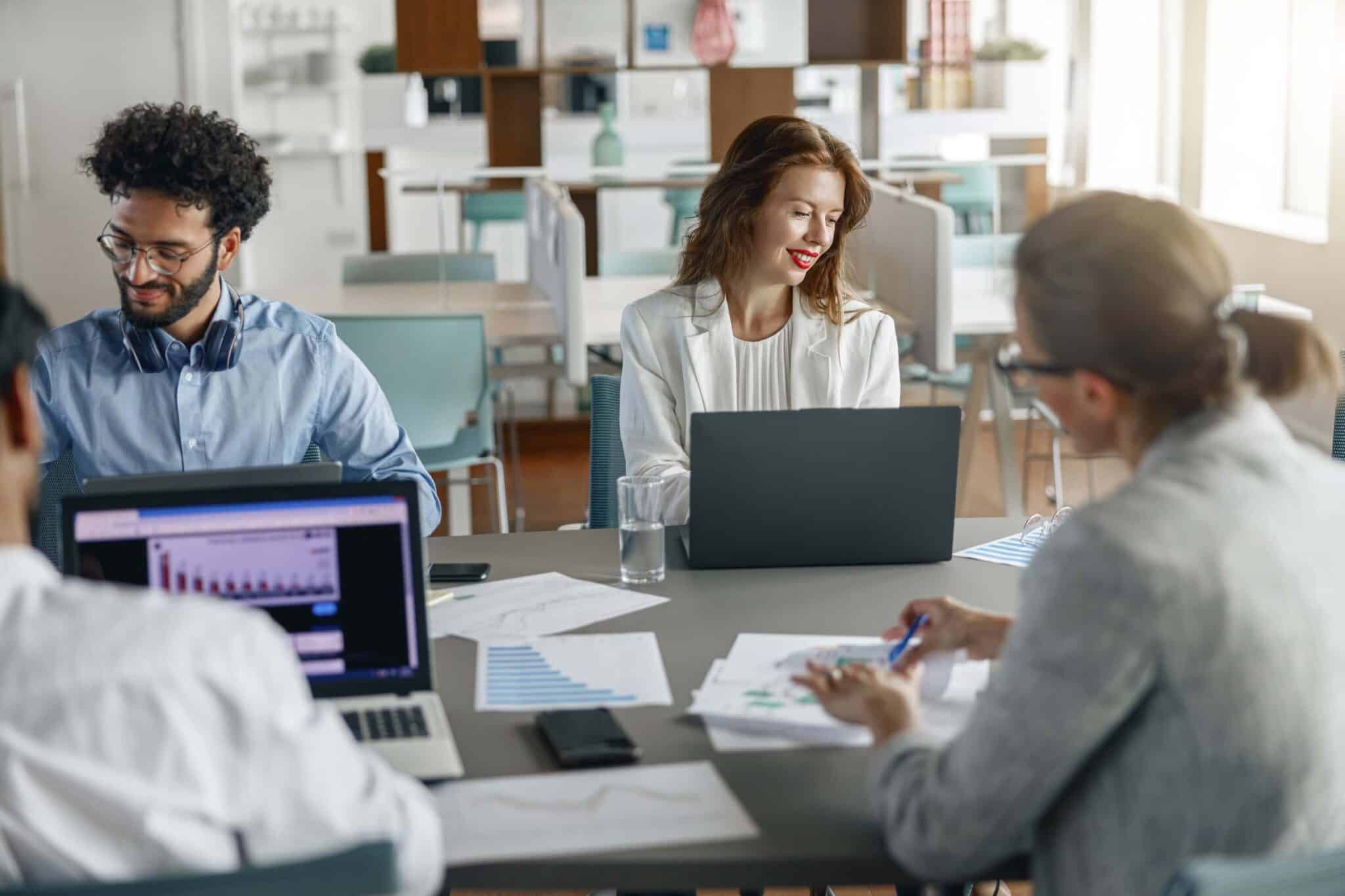 Four people sit around a table in a bright office space, working on laptops and reviewing charts and papers about manufacturing cash flow. A woman in white smiles at a colleague while others focus on their work.