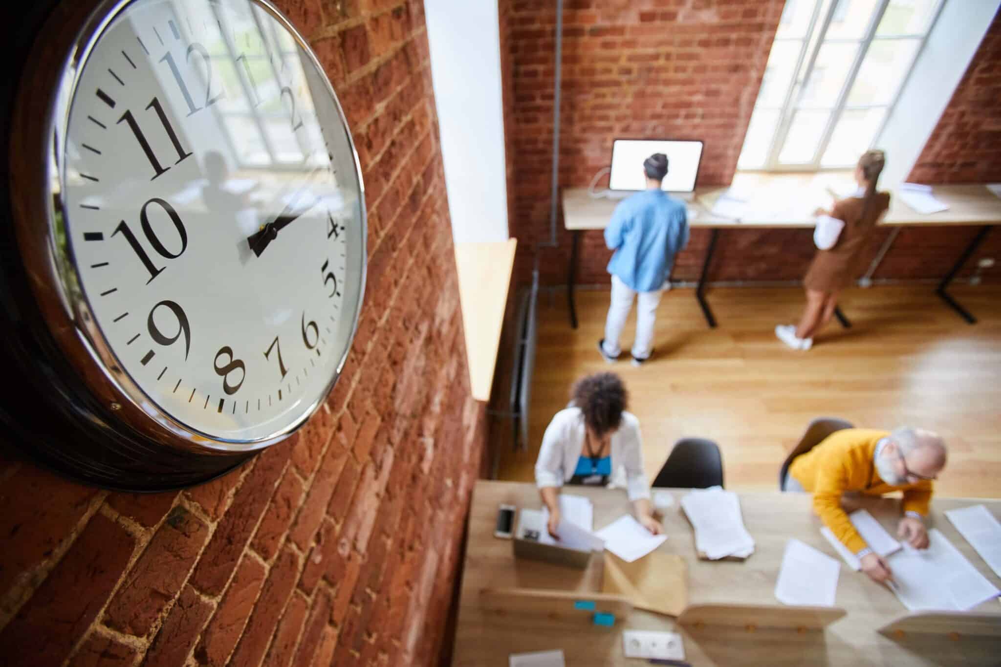 Large wall clock showing 9:08 hangs on a brick wall in a modern office space; four people are working at desks, some using computers and time tracking best practices, seen from an overhead angle.