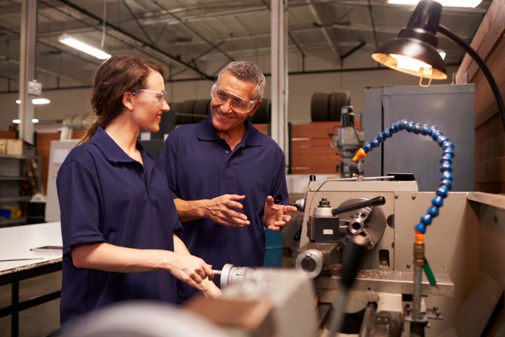 Two people wearing safety glasses and navy shirts talk and smile while working at a machine, showcasing efficient production management in a brightly lit factory. Industrial equipment and safety gear are visible in the background.