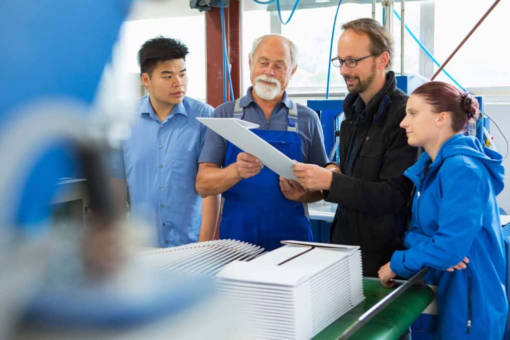 Four people in work uniforms stand around a table in a factory, reviewing a document together—demonstrating effective production management with equipment and stacks of papers or panels visible in the foreground.