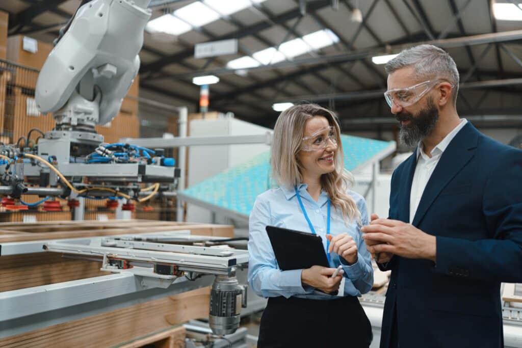 Two professionals in safety glasses and business attire stand in a modern factory, discussing production management near industrial robotic machinery. The woman holds a tablet and both are smiling, appearing engaged in conversation.
