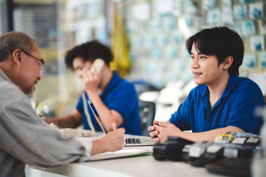 A young man in a blue shirt sits at a counter, attentively assisting an older customer who is filling out paperwork. Another employee behind them talks on the phone in a busy office environment.