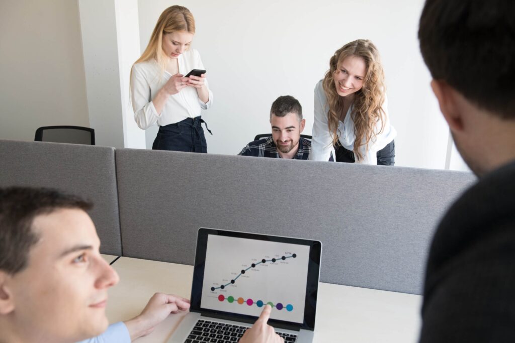 A group of young professionals in an office, with one person pointing at a laptop screen displaying a rising graph, while others stand and smile in the background.