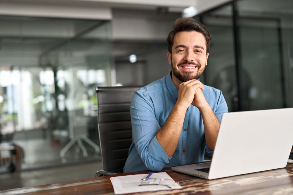A smiling man with a beard sits at a desk in an office, resting his chin on his hands. He is wearing a blue shirt and is positioned in front of a laptop, with paperwork and glasses on the desk.