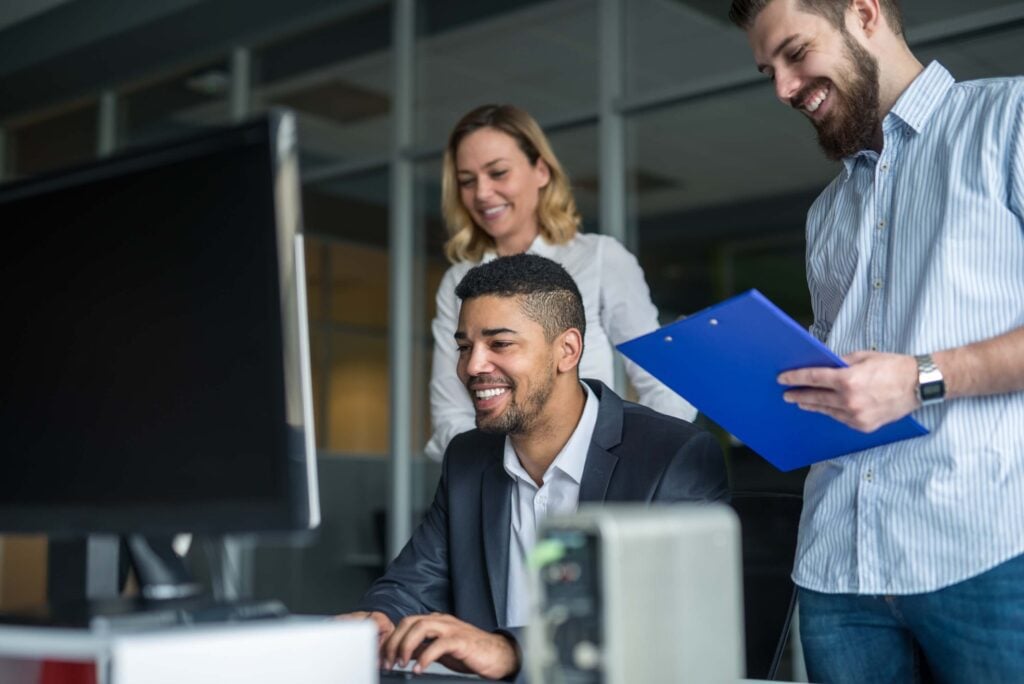 Three colleagues gather around a computer in an office, discussing sage production management. One man types and smiles at the screen, while a woman and another man with a clipboard stand nearby, all appearing engaged and cheerful.