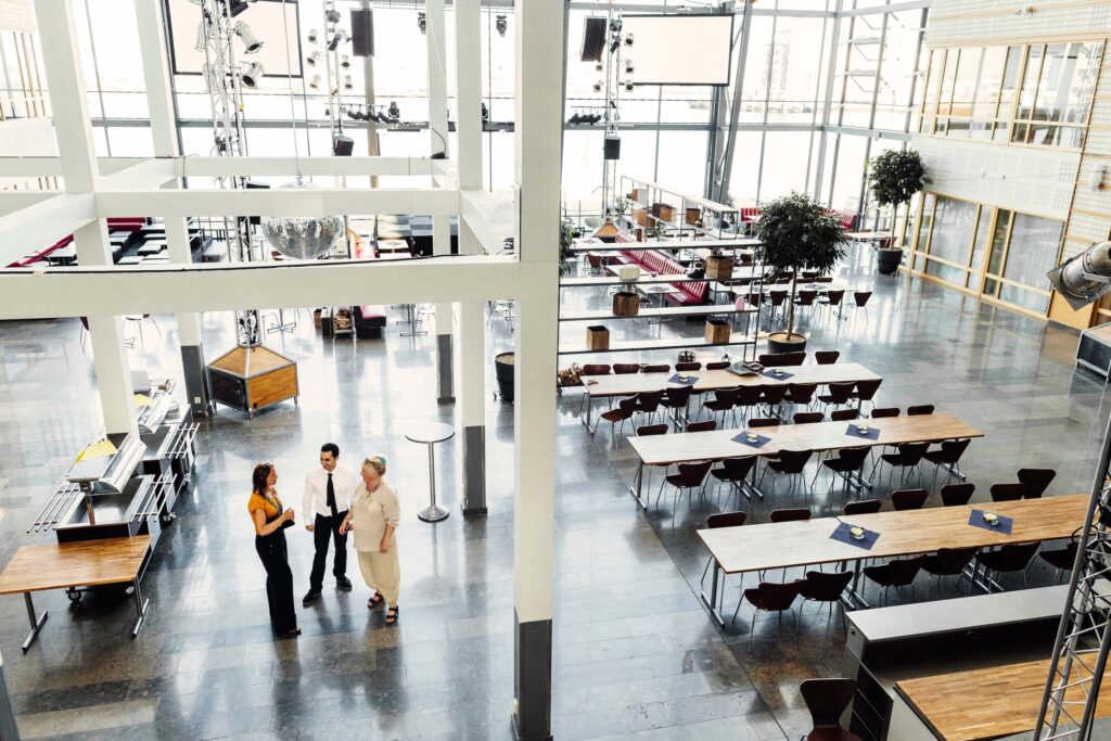 A spacious, modern atrium with large windows, wooden tables, and chairs. Three people stand and talk near the center, while the rest of the area appears empty and well-lit by natural light.