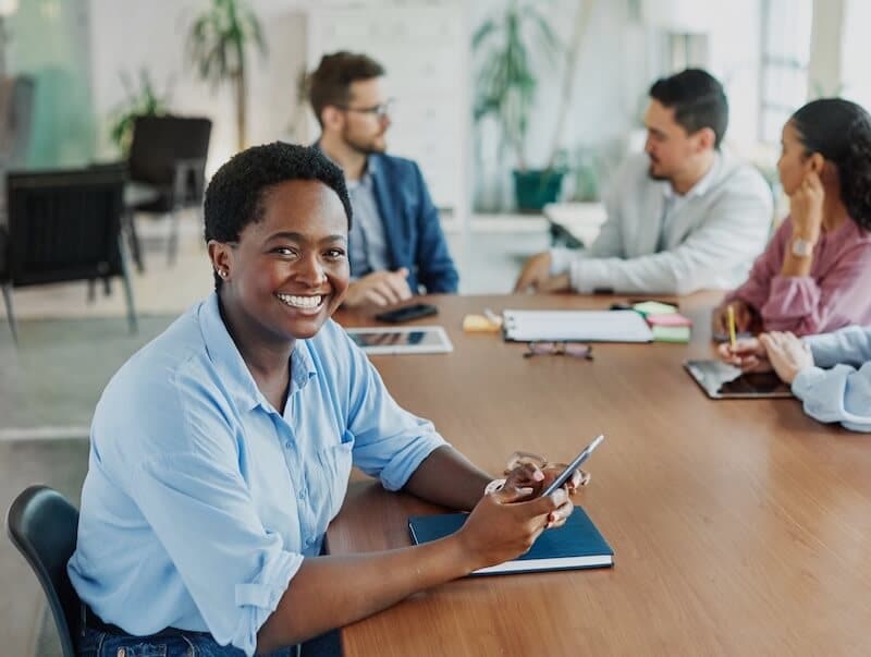 A smiling person sits at a conference table holding a phone, with three colleagues in the background engaged in discussion. The setting appears to be a modern, bright office space with plants and papers on the table.