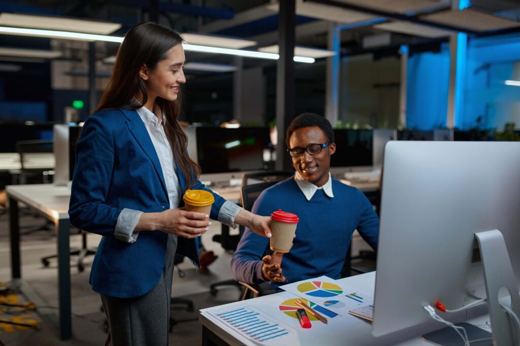 Two colleagues in a modern office smile as one hands a takeaway coffee to the other, who sits at a desk with charts, colored markers, and a computer monitor. The office has a relaxed, creative atmosphere.