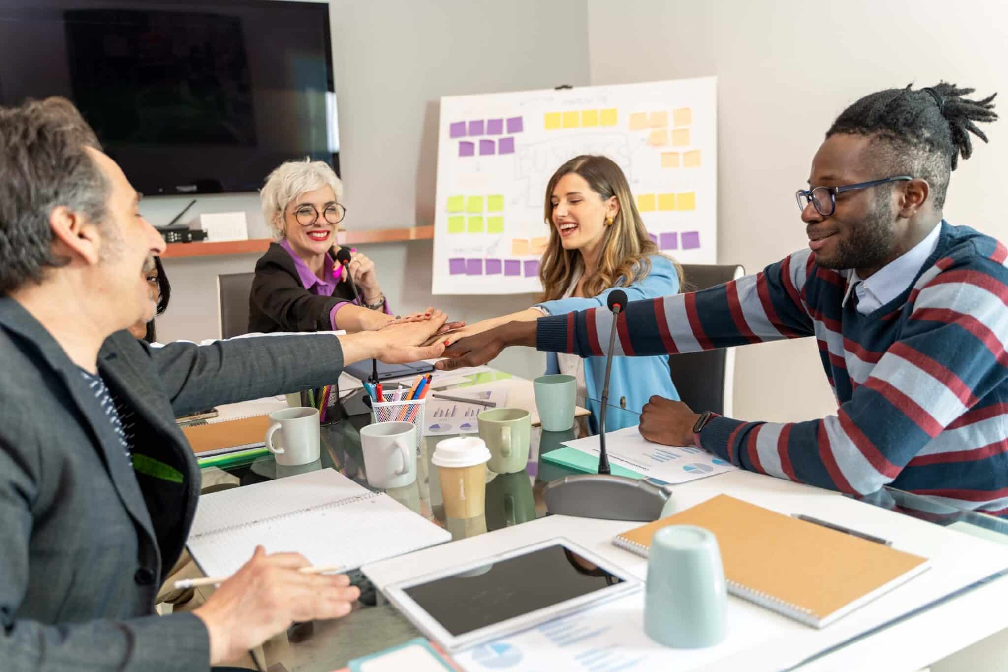 A diverse group of four people sit at a conference table discussing what is Replicon, smiling and stacking their hands together in a gesture of teamwork. Papers, notebooks, and mugs are on the table, with a colorful board in the background.