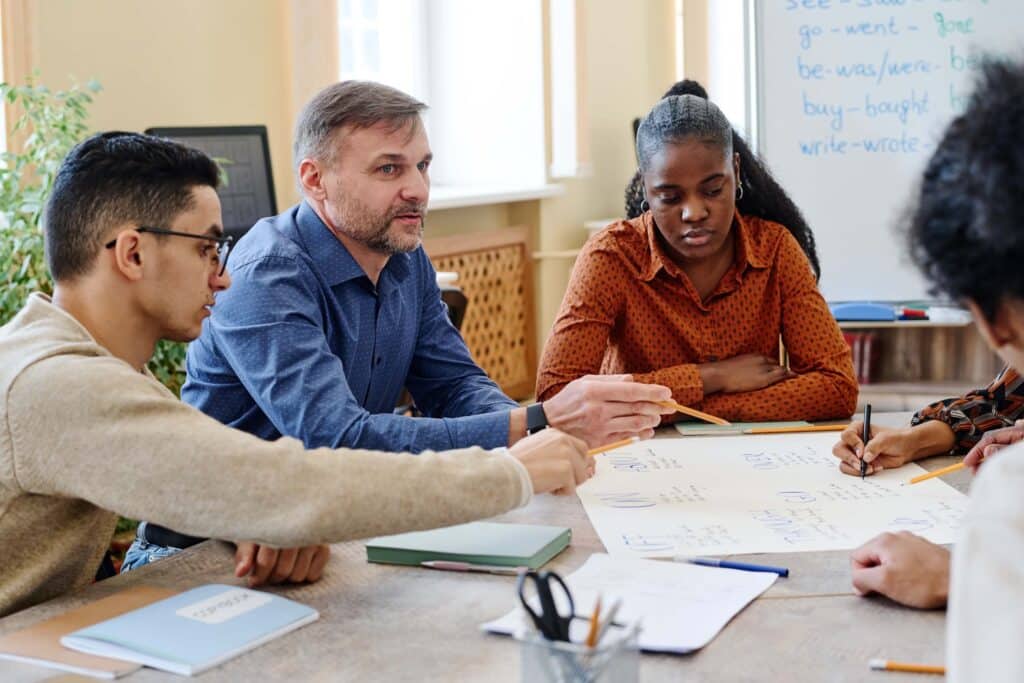 A group of four people sits around a table, collaborating and writing on a large sheet of paper. A whiteboard with notes is in the background, and notebooks and pens are on the table.