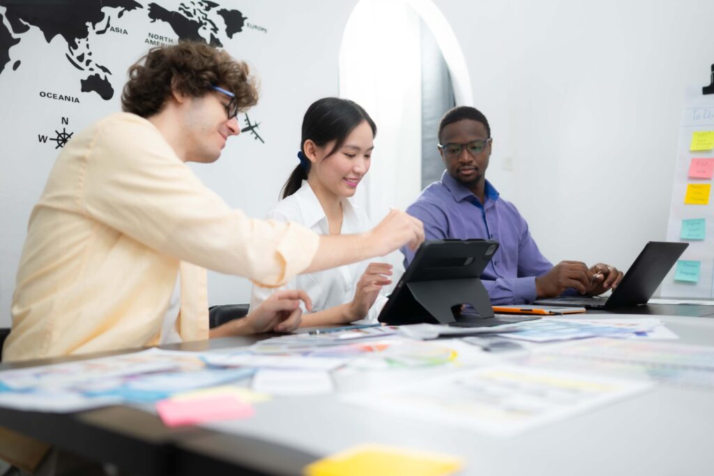 Three people sit at a desk collaborating, with laptops, tablets, and papers. A world map is on the wall behind them, and colorful sticky notes are visible on the side. All appear focused and engaged in discussion.