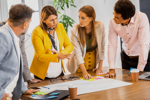 Four colleagues stand around a table covered with papers and sticky notes, engaged in a serious discussion. A woman in a yellow blazer gestures while speaking, and the others listen attentively.