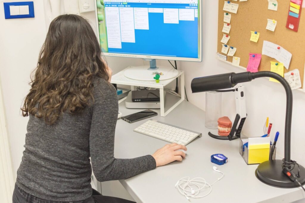 A person with long dark hair sits at a desk, looking at a computer monitor with a task board displayed. The desk has office supplies, a lamp, dental model, and a corkboard with colorful sticky notes in the background.