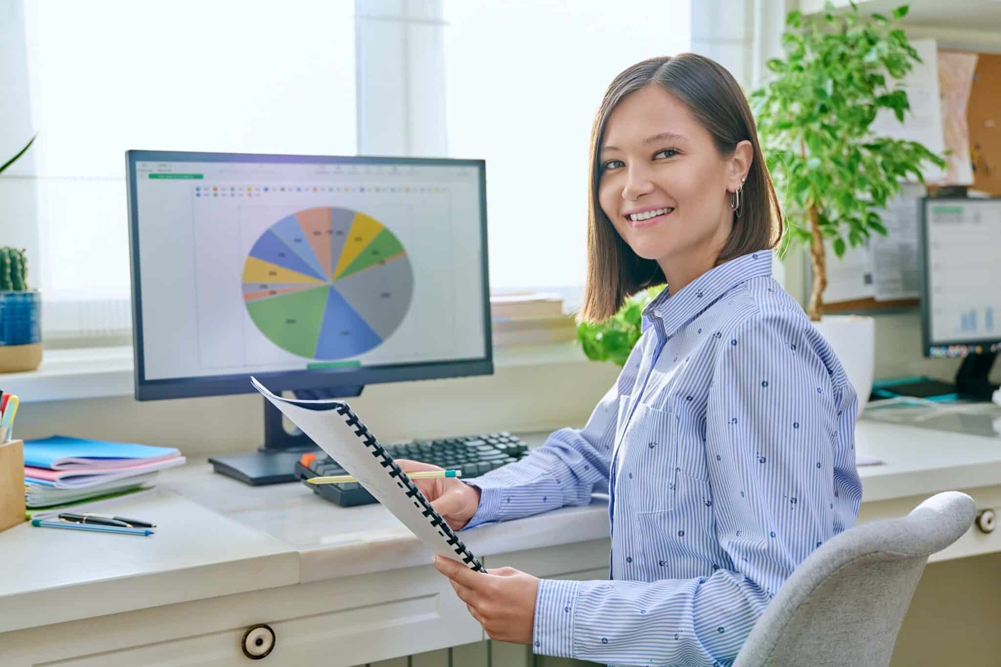 A woman sits at a desk in a bright office, smiling at the camera. She holds a report and faces a computer monitor displaying a colorful pie chart about employee utilization. The workspace has potted plants, papers, and office supplies.