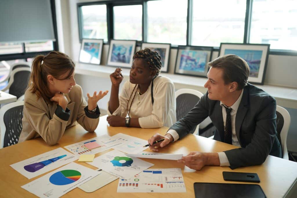 Three business professionals sit at a conference table discussing charts and graphs, analyzing data with cost management software. Papers, charts, and a tablet are spread on the table. Framed pictures and windows are visible in the background.