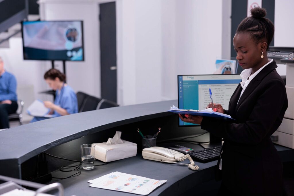 A receptionist in a black suit writes on a clipboard at a modern medical office front desk. A computer, phone, tissue box, and documents are on the counter. Two people are seated in the waiting area in the background.