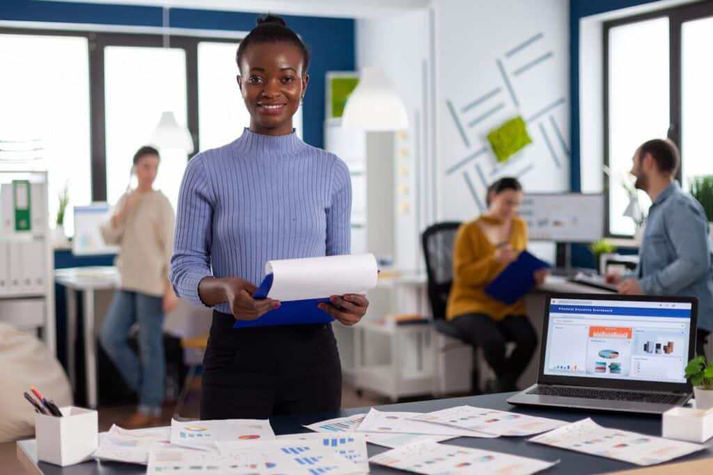 A woman stands at a desk holding papers, smiling at the camera in a modern office. The desk is covered with charts and documents, and a laptop displays graphs. Three colleagues work in the background.