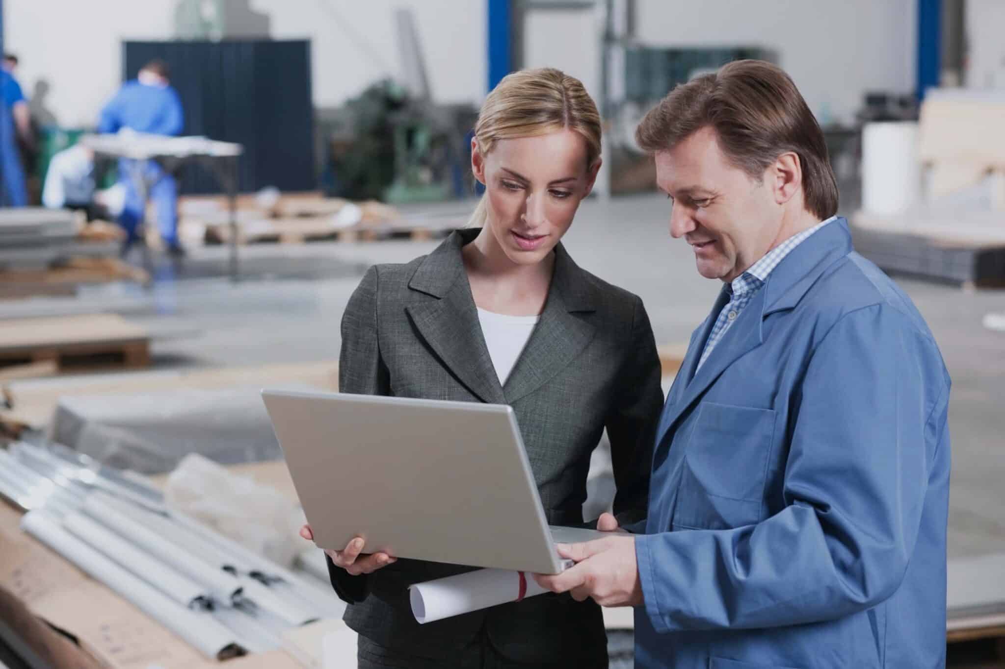 A woman in a business suit and a man in a blue lab coat review sage 100 production management data on a laptop together in an industrial warehouse, with materials and workers visible in the background.