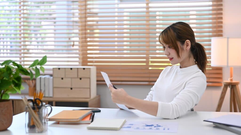 A woman sits at a desk in a bright office, holding and reading a sheet of paper. The desk has books, a potted plant, and office supplies. Blinds cover the window in the background, letting in natural light.