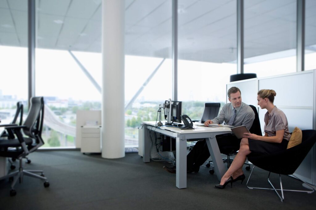 A man and a woman in business attire sit in a modern office by large windows, discussing ERP Timesheet documents at a desk with computers, while city buildings are visible outside.