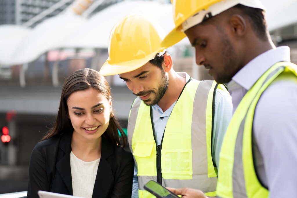 Three construction professionals, two men in yellow safety vests and hard hats and a woman in business attire, collaborate outdoors at a work site, reviewing project data on a tablet to ensure DCAA compliant time tracking.