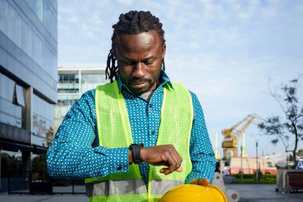 A construction worker wearing a green safety vest and blue shirt looks at his smartwatch while holding a yellow hard hat outdoors, with cranes and buildings in the background.
