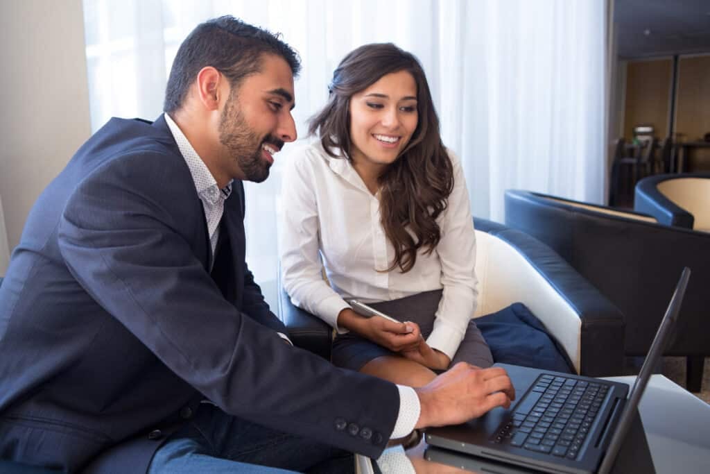 A man in a suit and a woman in business attire sit together, smiling and looking at a laptop screen in a modern office setting, reviewing timesheet compliance for hourly and salaried employees.