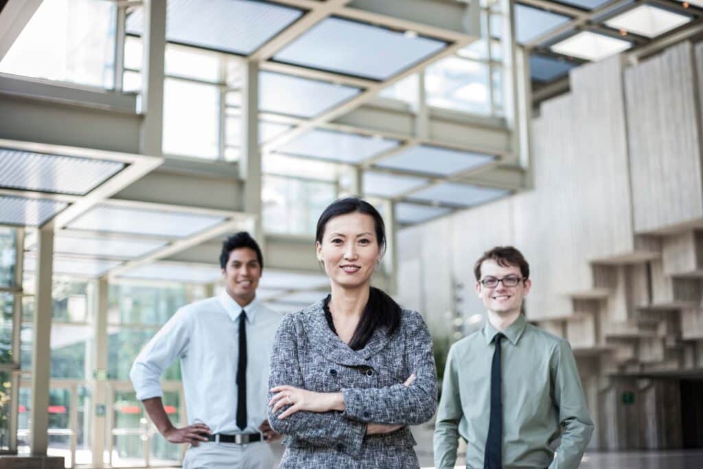 Three professionally dressed people stand confidently in a modern, glass-walled office atrium, embodying teamwork and effective workforce management. A woman stands in front with arms crossed, while two men smile behind her, all facing the camera.