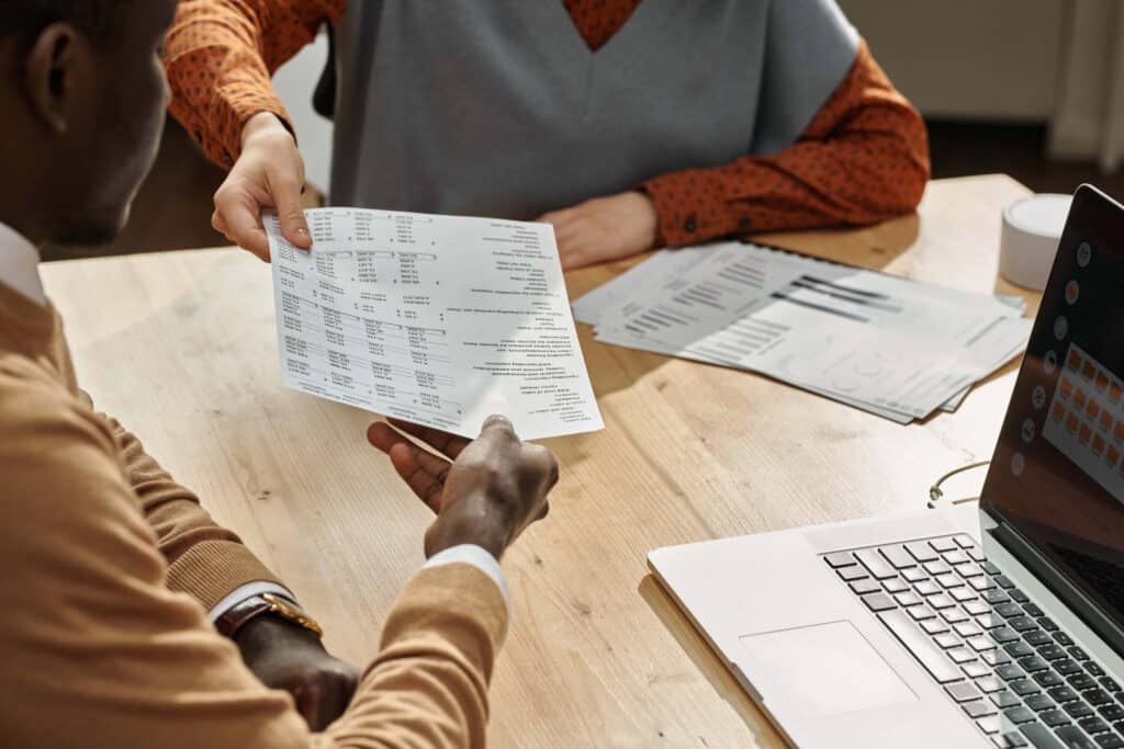 Two project managers sit at a wooden desk with a laptop, reviewing an ERP Timesheet printed with tables and numbers. One hands the sheet to the other, while additional papers are scattered on the desk.
