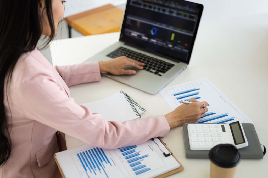 A woman in a pink blazer works at a desk with a laptop, writing in a notebook. Papers with bar charts and an ERP Timesheet, plus a calculator and coffee cup, are on the table. Data is displayed on the laptop screen—ideal for project managers.