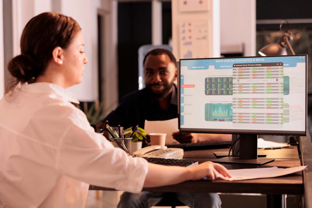 Two people sit at a desk in an organized office. One woman uses a computer displaying ERP Timesheet data and graphs. The man in the background, likely one of the project managers, holds papers and smiles among plants and office supplies.