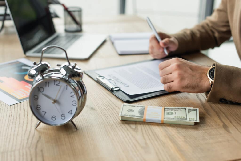 A person in business attire writes on a clipboard at a desk with $100 bills, an alarm clock, and documents—highlighting financial or business work, possibly related to timesheet compliance for hourly and salaried employees.