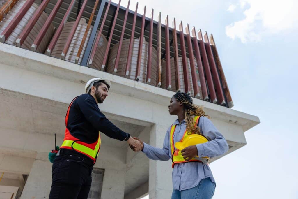 Two construction workers wearing safety vests and helmets shake hands at a building site, with one holding a yellow hard hat and blueprints, standing in front of a concrete structure, highlighting the importance of DCAA compliant time tracking.