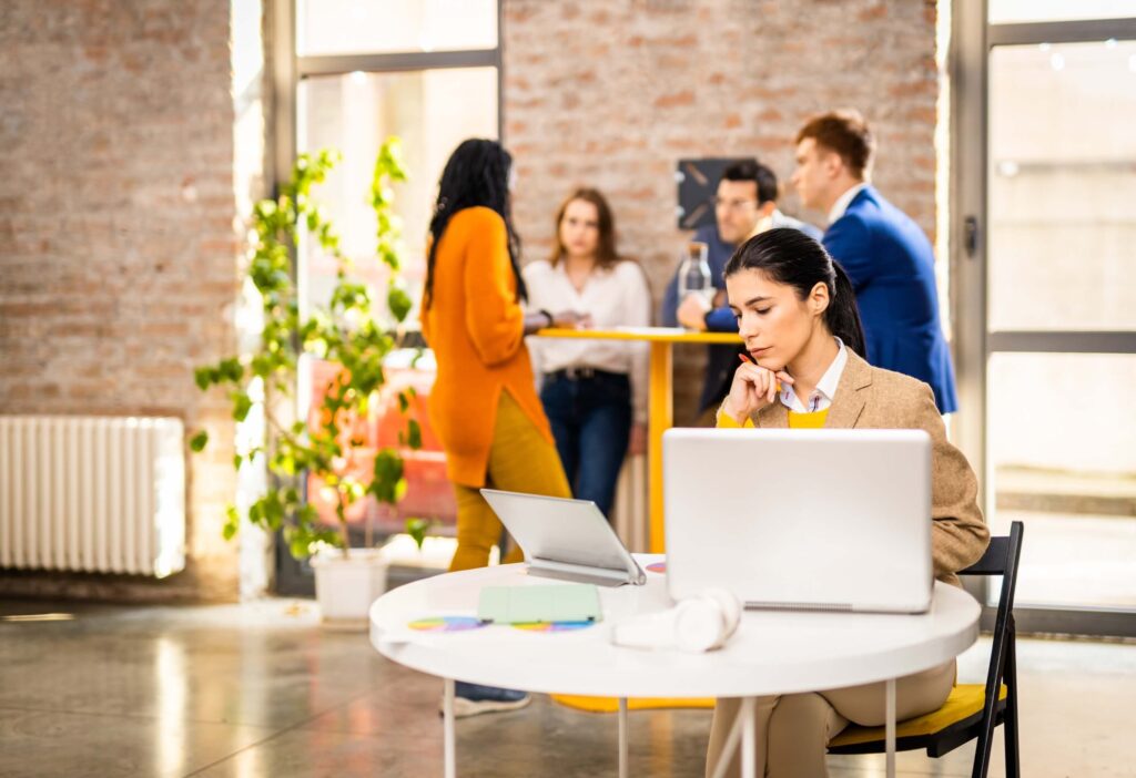 A woman sits at a round table with two laptops, concentrating on her work—perhaps using AI time tracking to boost productivity. In the background, four people stand and talk near a high table in a bright office with large windows and exposed brick walls.