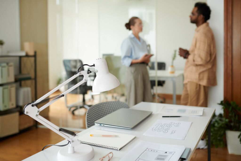 A modern office desk with a lamp, laptop, notebooks, and papers in the foreground. In the background, two people stand and talk near glass walls and shelves. The scene is bright and organized, perfect for using AI time tracking software.