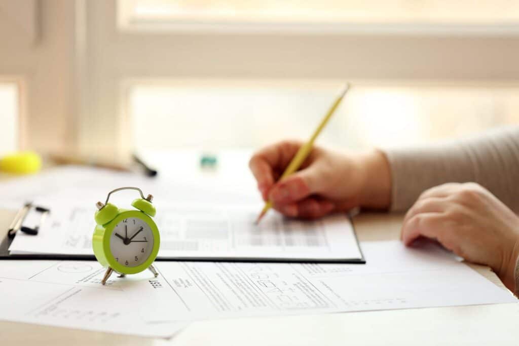 A small green alarm clock sits on a desk next to paperwork, while a person writes on a clipboard with a yellow pencil—creating an organized workspace ideal for project management. The background shows a sunlit window.
