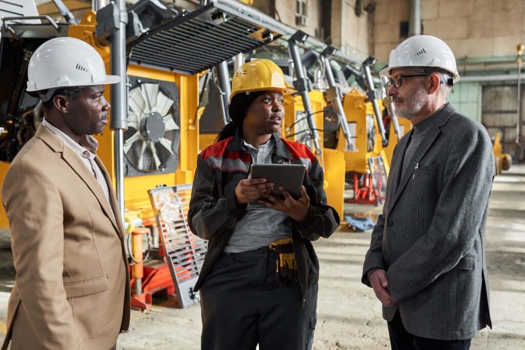 Three people wearing safety helmets stand in an industrial setting with machinery in the background. The person in the center holds a tablet, possibly discussing DCAA compliant time tracking, while the others listen attentively.