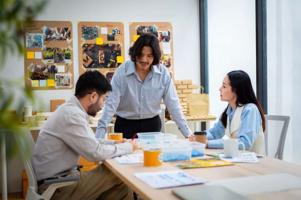 Three people are having a discussion at a table in a brightly lit office, reviewing papers and coffee cups. Display boards with photos and notes highlight their focus on project management and effective workforce management strategies.
