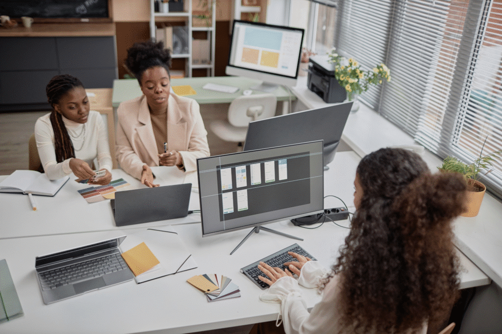 Three women in an office collaborate at a desk with laptops, notebooks, color swatches, and a desktop monitor displaying a design layout. They discuss timesheet compliance for hourly and salaried employees as sunlight streams through large windows.