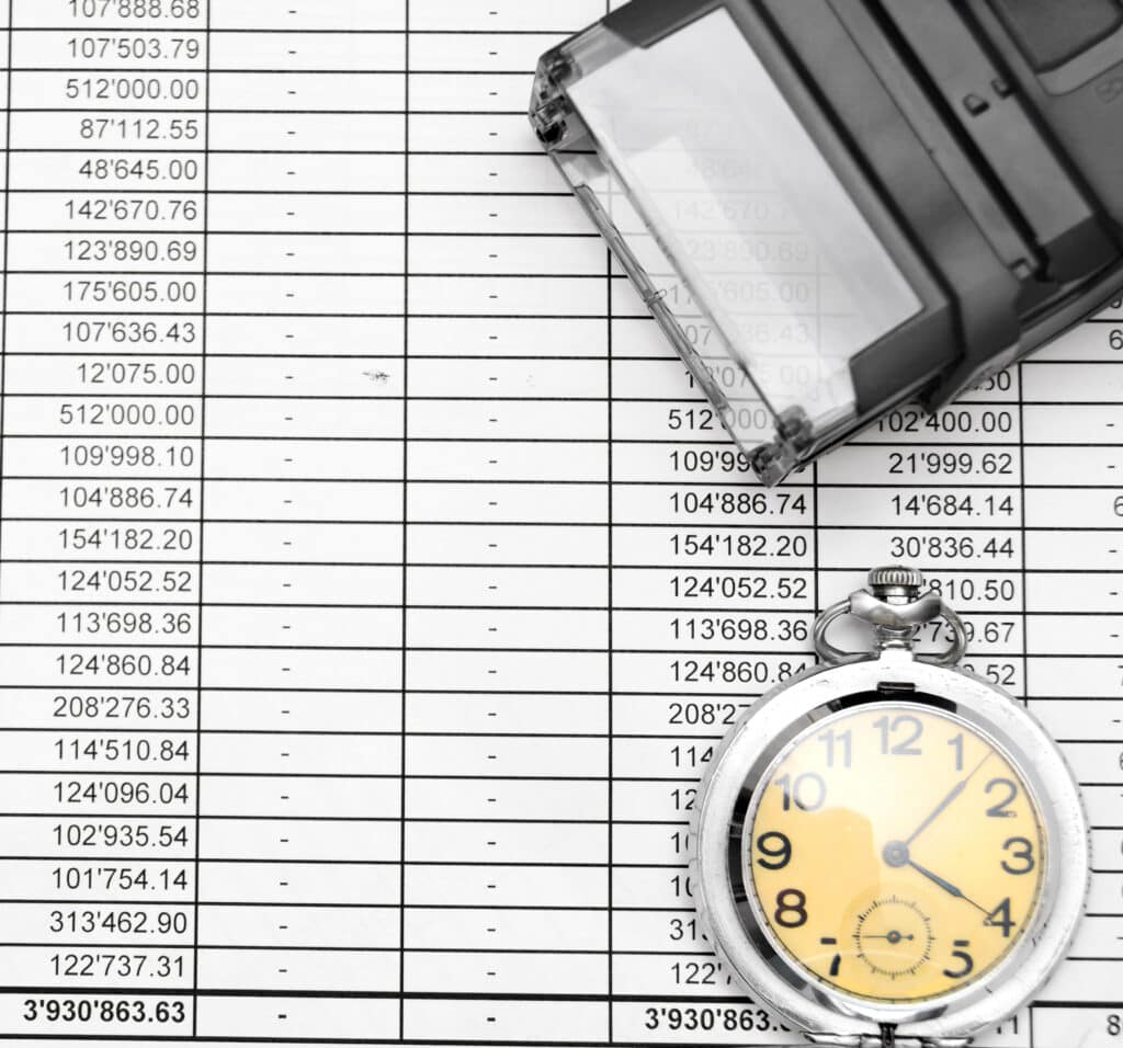 A black and white calculator and a silver pocket watch lie on top of a printed spreadsheet filled with numerical data, highlighting the importance of timesheet compliance for hourly and salaried employees.
